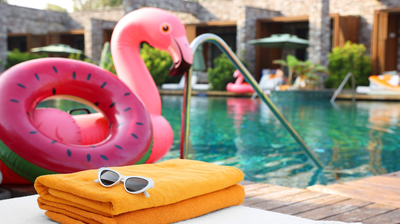 A folded yellow towel and sunglasses next to pink pool floats and a pool.