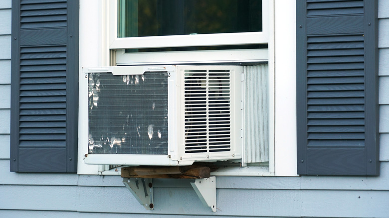 An old air conditioner sits in the window of a house with blue side panels and dark blue shutters