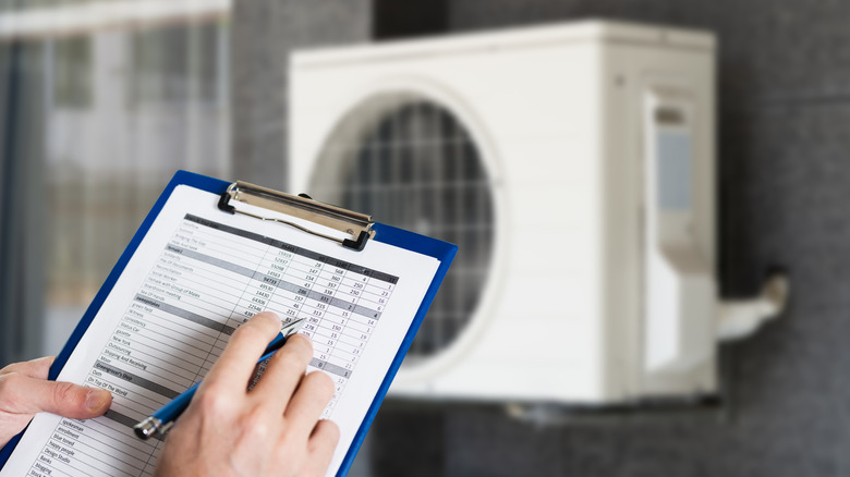 A repair technician holds paperwork on a clip board with a blurred air conditioner in the background