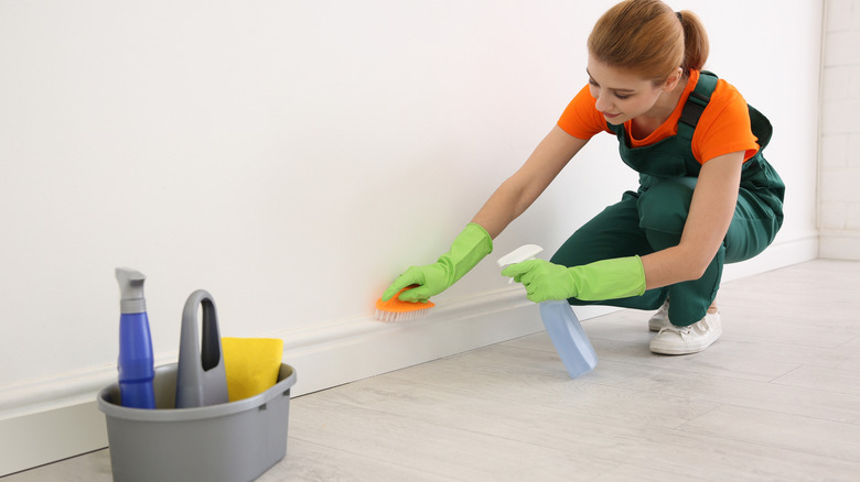 Woman kneeling down to clean baseboards with spray and scrub brush