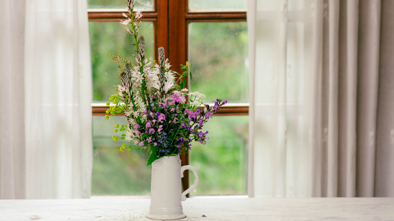 Colorful wildflowers arranged in a white beer stein, placed on a wooden table near a window with soft, sheer curtains