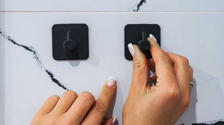 A person mounts a second black plastic hook onto a marble tiled bathroom wall.