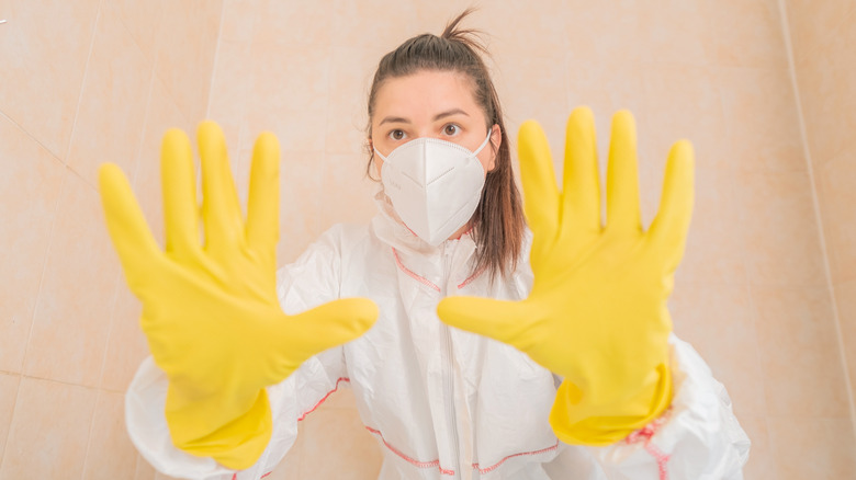 A woman cleaning the bathroom in protective gear shows her gloved hands to the camera.