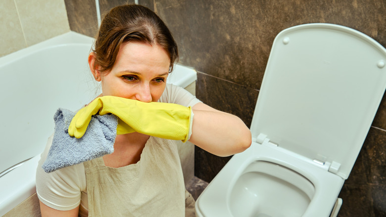 A woman cleaning the home bathroom with a sense of disgust while sitting near the toilet bowl.