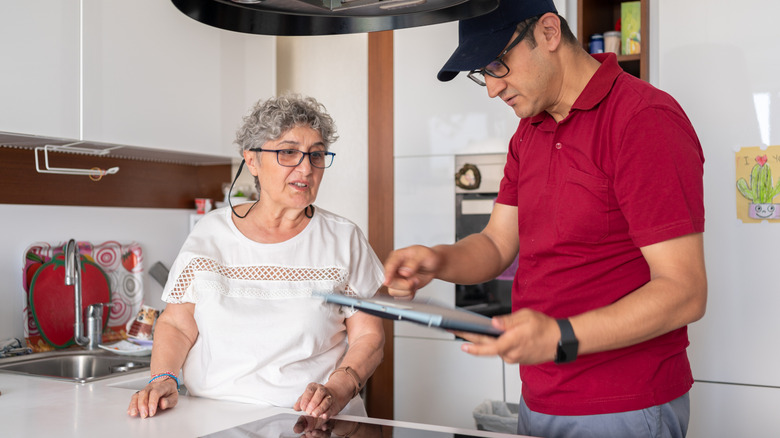 A repair man pointing at a clipboard next to an elderly woman in her kitchen