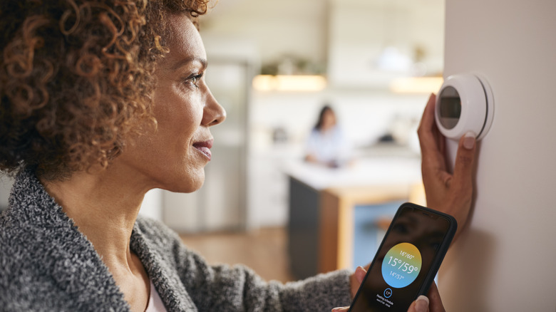 Woman Using App On Phone To Control Digital Central Heating Thermostat At Home