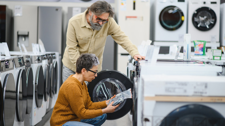 A couple in an appliance store compare washing machines. The woman is wearing an orange sweater and jeans, and crouches on the ground, while the man stands over her looking at the product.