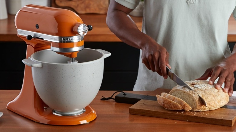 Someone cuts a loaf of fresh bread next to an orange KitchenAid stand mixer
