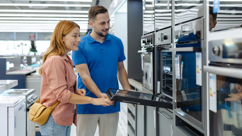 A young couple look at different models of wall ovens inside of a store.