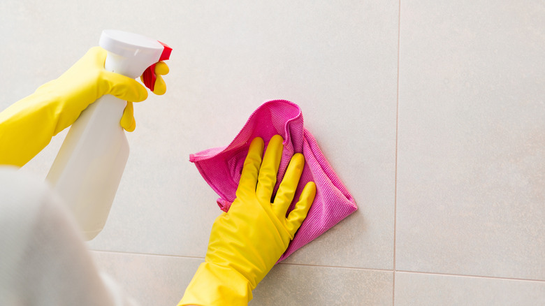 Hands with yellow rubber gloves holding detergent spray bottle and cleaning tiles in bathroom with pink cloth.