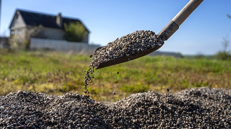 Close-up of a shovel full of gravel as it pours it onto a pile of gravel with a blurred grass background