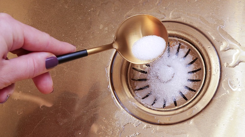 A person's hand with purple nail polish pours a spoon of a white substance down the sink drain