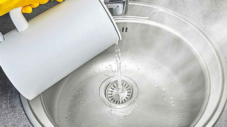 A gloved hand is pouring boiling water down a sink drain from a kettle