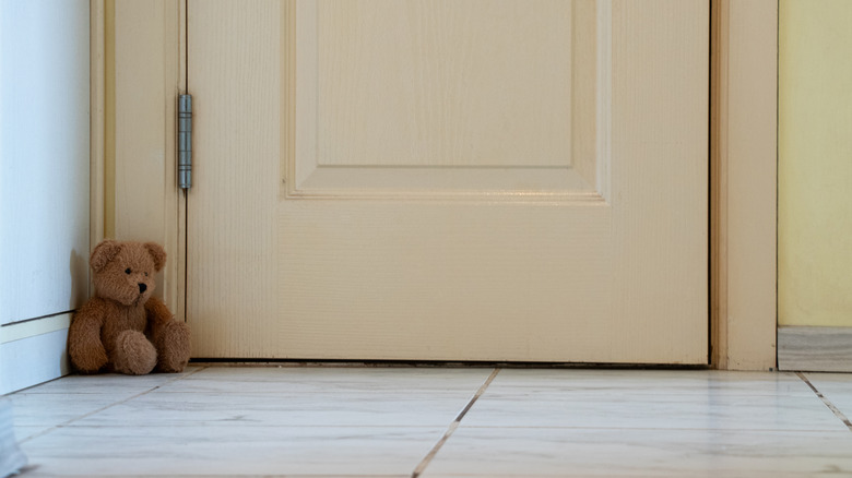 A beige door with a visible gap along the edge, set against a tiled floor, with a brown teddy bear tucked in the corner