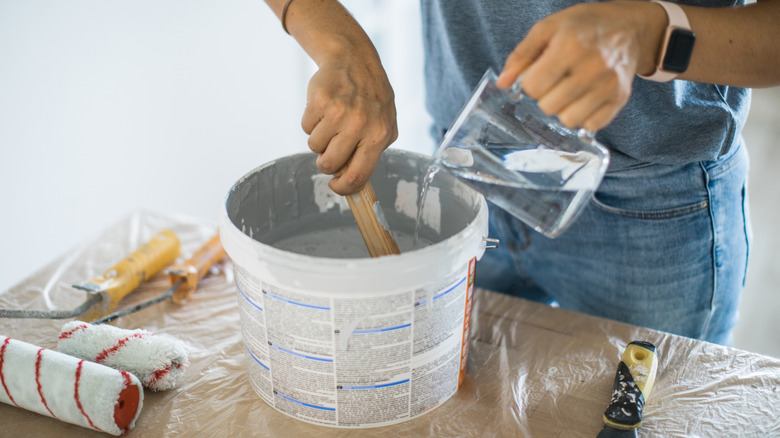A person mixing water into gray paint on top of a plastic sheet with new rollers on the side