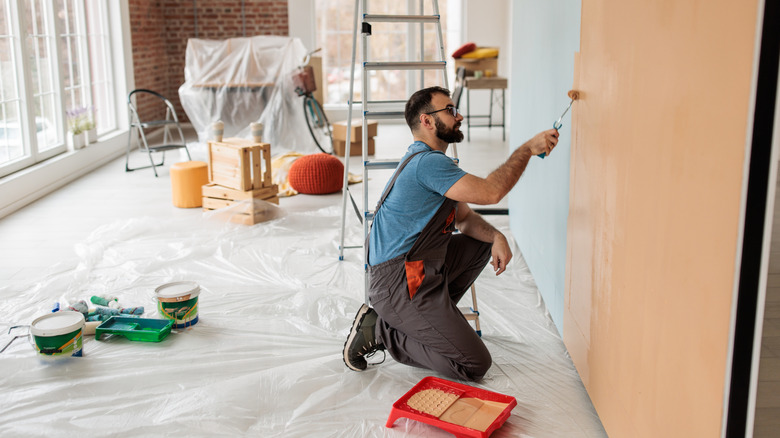 A person kneeling down on a plastic-covered floor, using a roller to paint the wall orange