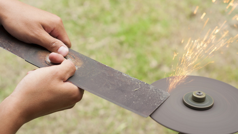 Two hands hold a lawn mower blade against a rotating sharpener outdoors.