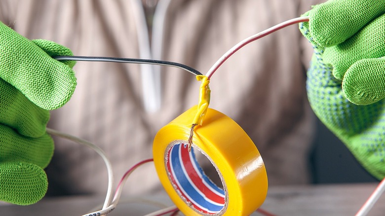 Close-up of a person wearing green gloves holding wires wrapped in yellow electrical tape