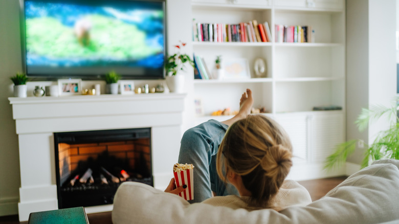 A blonde woman eating popcorn on a couch while watching a TV placed above a lit fireplace