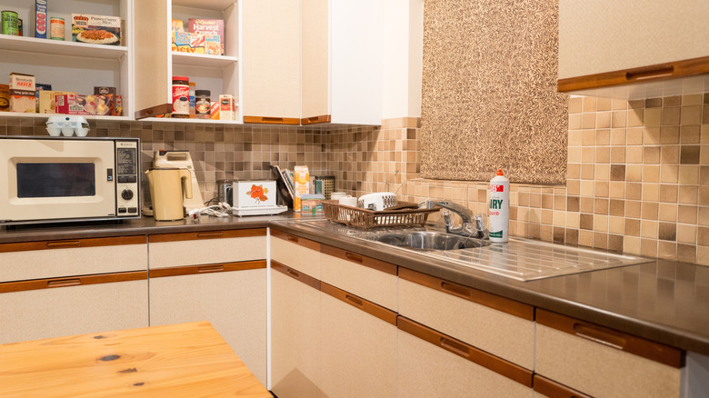 An outdated kitchen with cream cupboards and brown trim and a backsplash with small square tiles in brown, beige and cream tones