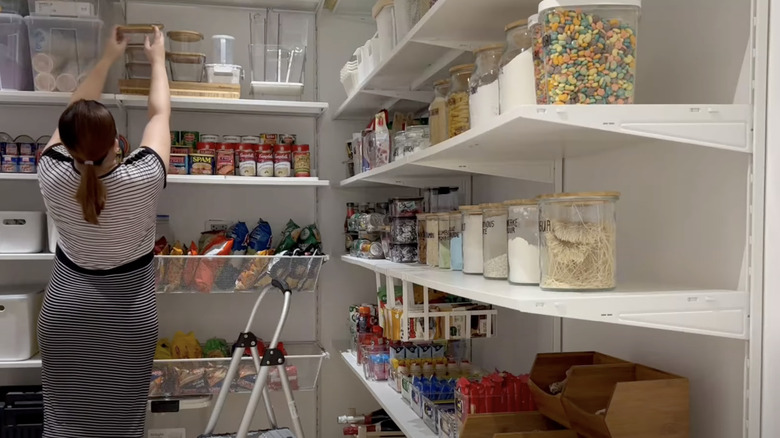 A person adds food storage containers to the top shelf of her pantry customized with IKEA BOAXEL products