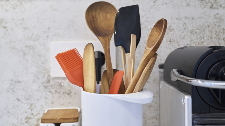 Close-up of front view of different kitchen utensils inside white container against a textured wall on worktop