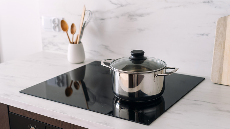Selective focus on stainless steel pan with lid at glass ceramic hob against marble kitchen table on blurred background. Cooking food at home on modern induction stove. Metal kitchenware on cooker