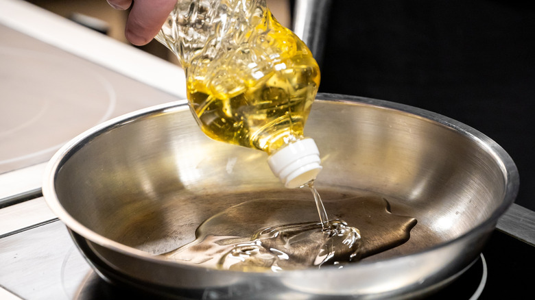 A hand pours golden cooking oil from a plastic bottle into a stainless steel frying pan on a stovetop.
