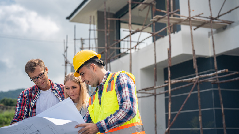 Two people look at construction plans with a construction worker in front of a house.