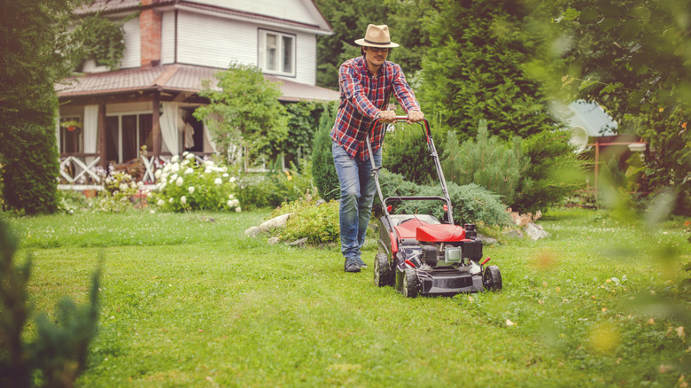 A person wearing a sun hate, jeans, and a long-sleeved shirt mowing their grass