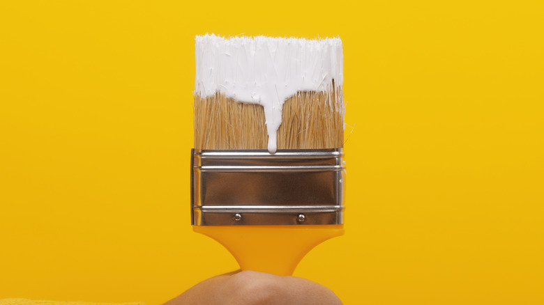 A paintbrush with white paint being held in front of a yellow wall.