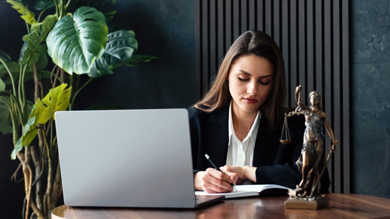 A female lawyer sitting at a desk and writing in a notebook with a plant in the corner.