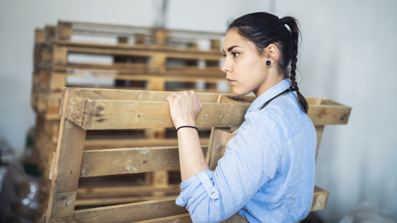 Young woman holding a wood pallet with more pallets stacked in the background
