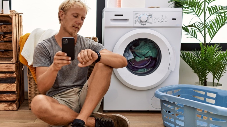 man checking watch and holding smartphone while sitting on floor waiting for washing machine to finish