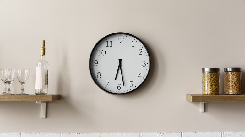 A standard black-and-white clock hanging on an off-white colored wall with shelves holding kitchen goods on either side.