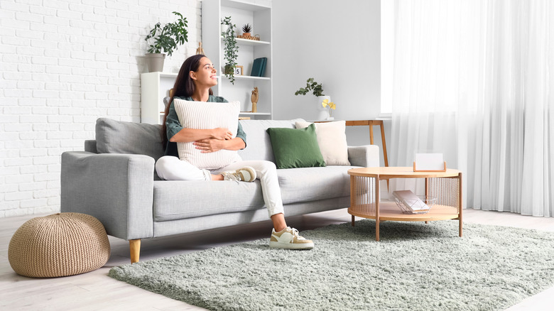 A woman sitting on a grey couch holding a white pillow with a wooden coffee table off to one side of her and a tan pouf on the other