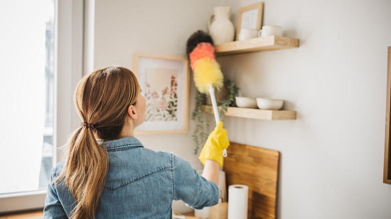woman in yellow gloves dusting wooden open shelves with white ceramic bowls on them with a fluffy black, pink, and yellow duster