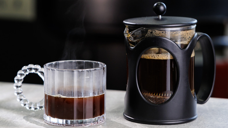 French press with fresh coffee in it near a half filled glass coffee mug on a counter