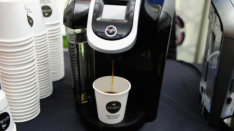 Keurig coffee machine dispensing coffee into a disposable cup with large stacks of cups near it.