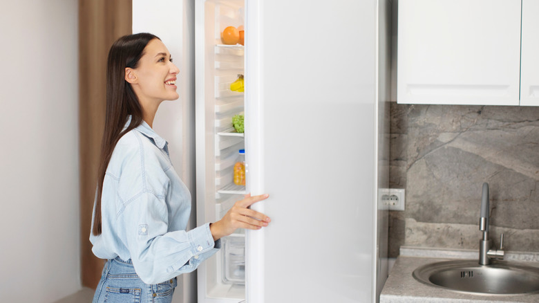 A woman opening a white fridge and smiling as she looks at the shelves in the door