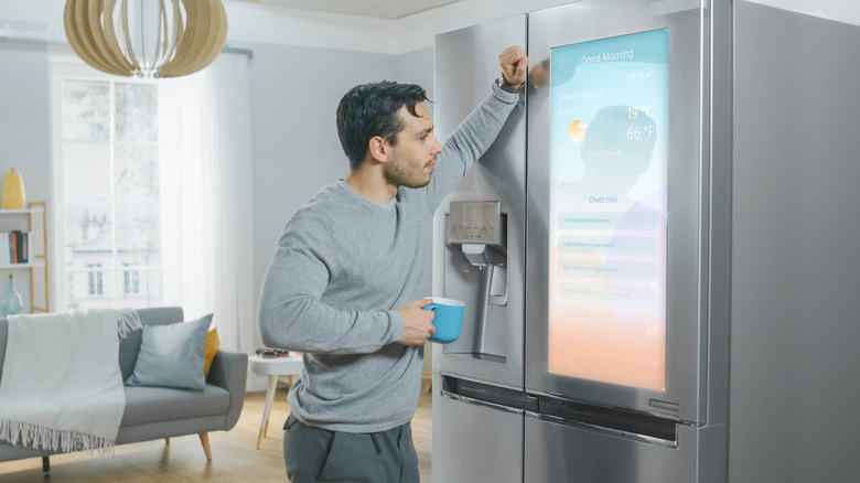 A man leaning on a fridge with a cup of coffee, looking at a smart fridge screen