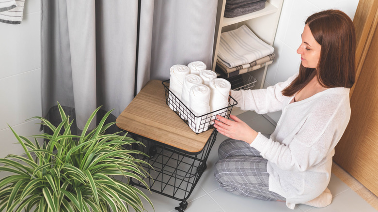 A woman puts a wire basket full of rolled white towels on a small wheeled side table in a bathroom.