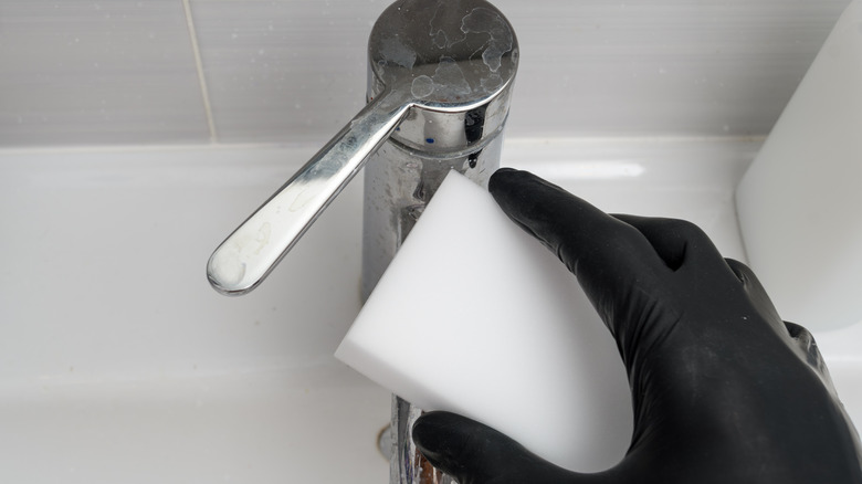 Hand in a black glove using a Magic Eraser to clean a bathroom faucet
