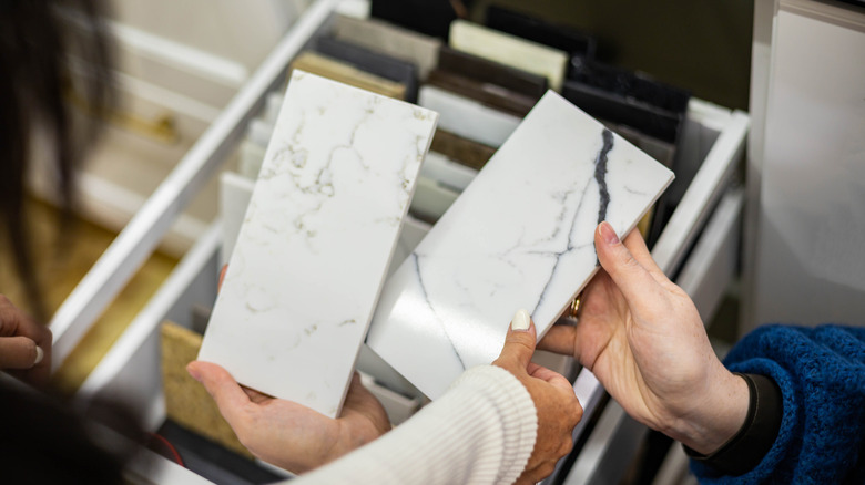 Two pairs of hands are holding up two rectangular samples of stone, while various colors of samples are in a blurred drawer below.