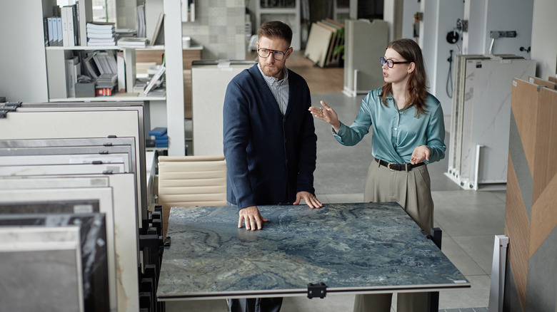 A man wearing glasses and a long-sleeved blue cardigan and a woman with glasses and a silky teal shirt are standing next to a stone slab, surrounded by other countertop samples.