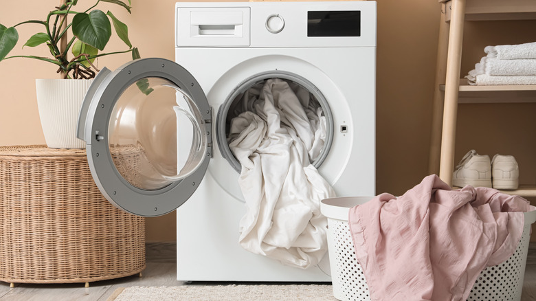 A laundry basket with pink bedsheets sits in front of a white washer loaded with white sheets.