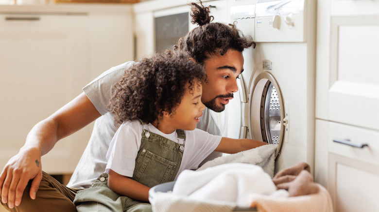 Father and child put clothes into a washing machine together.