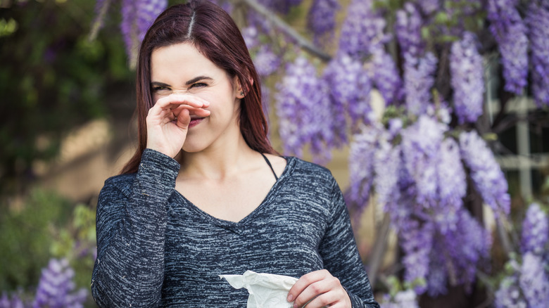 A woman covering her nose in a garden with purple hanging flowers behind her