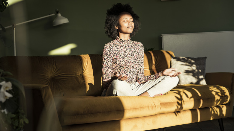 Woman meditating on dark yellow couch in green-painted room.