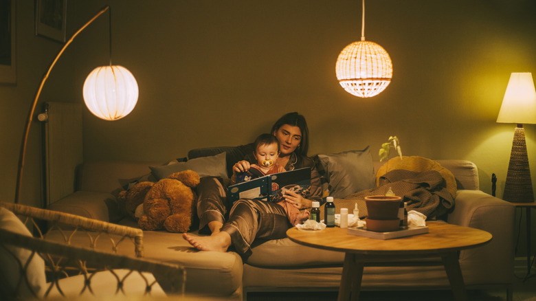 A mother reads her baby a story on a comfortable couch in a room with warm lighting from three different lamps.